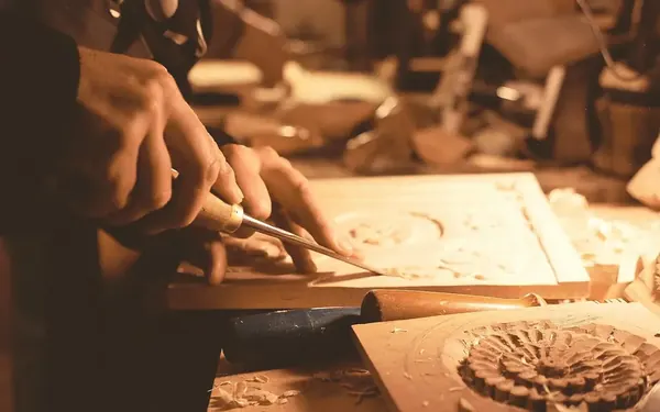 A woodcarver's hands cutting a pattern into a panel with a chisel