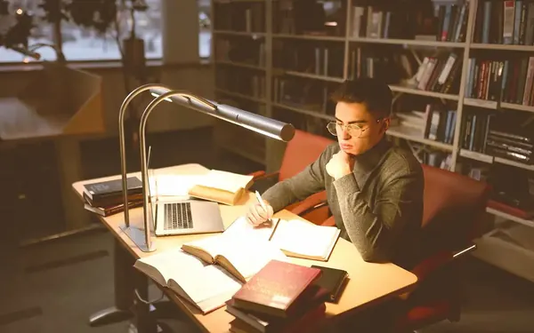 A student working under a desk lamp at a library table stacked with books